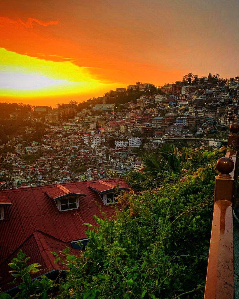 Golden sunset view over the hillside city of Shimla, featuring the colonial red tin roof of Clarkes Hotel in the foreground and the city layers in the background.