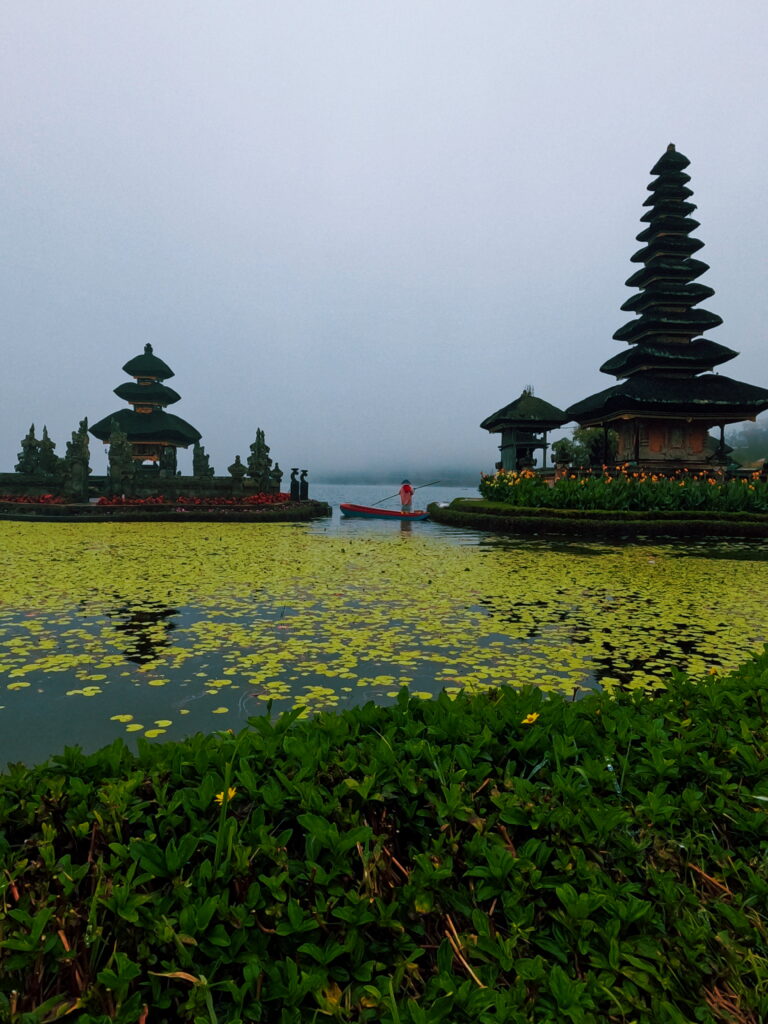 A misty morning view of the Pura Ulun Danu Bratan Hindu temple on Lake Bratan, Bali. A small boat is on the water surrounded by green lily pads and the temple's multi-tiered meru towers.